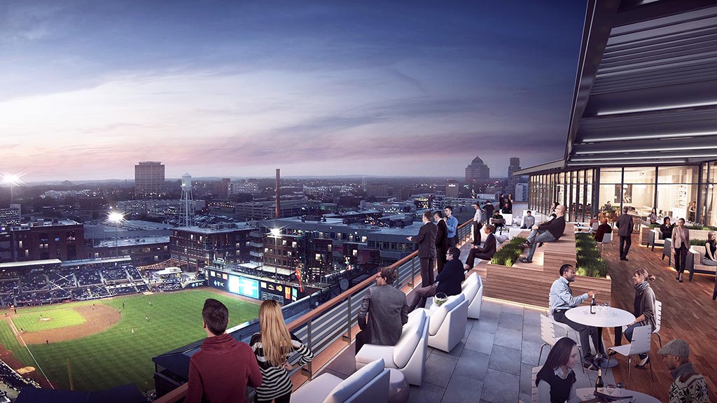 A group of people sitting at tables in a large open area with a large grassy field and buildings.