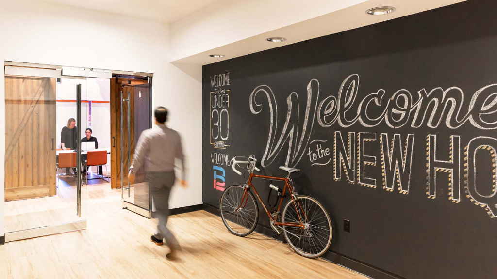 A person walking in a room with a bicycle and a chalkboard.