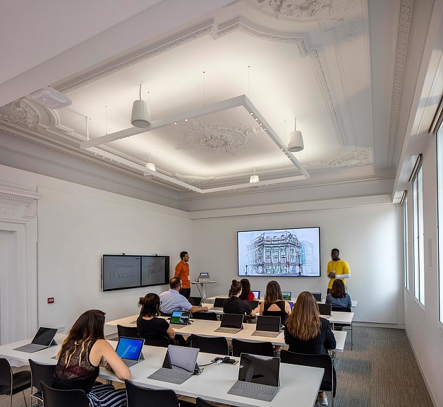 A group of people sitting at tables with laptops in a room with a large screen.