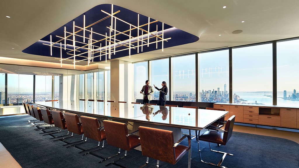 A couple of people standing in a room with large windows and a table.