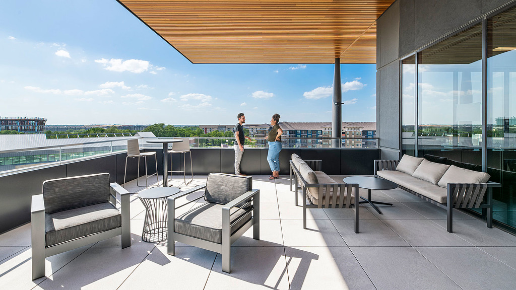 A couple of people standing next to a building with benches and a body of water in the background.