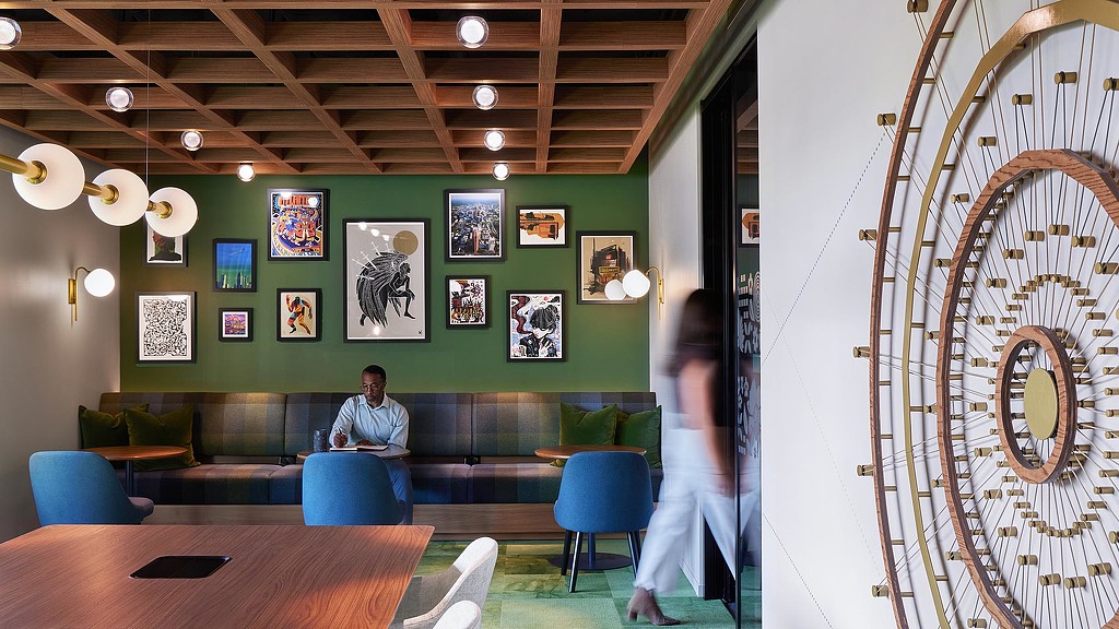 A person sitting in a room with a green wall and a person standing in front of a table.