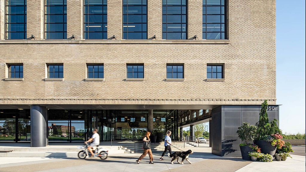 People walking a dog and riding a bike outside of a large building with a brick facade