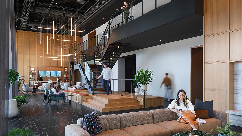 A person sitting on a couch in a room with people walking and a staircase.