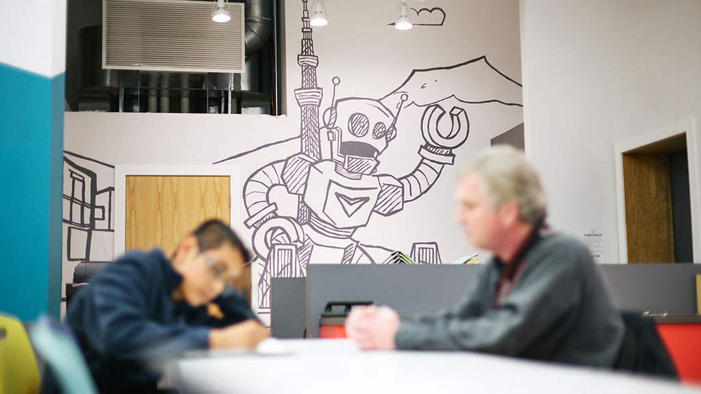 A man and a woman sitting at a table with a drawing on the wall.