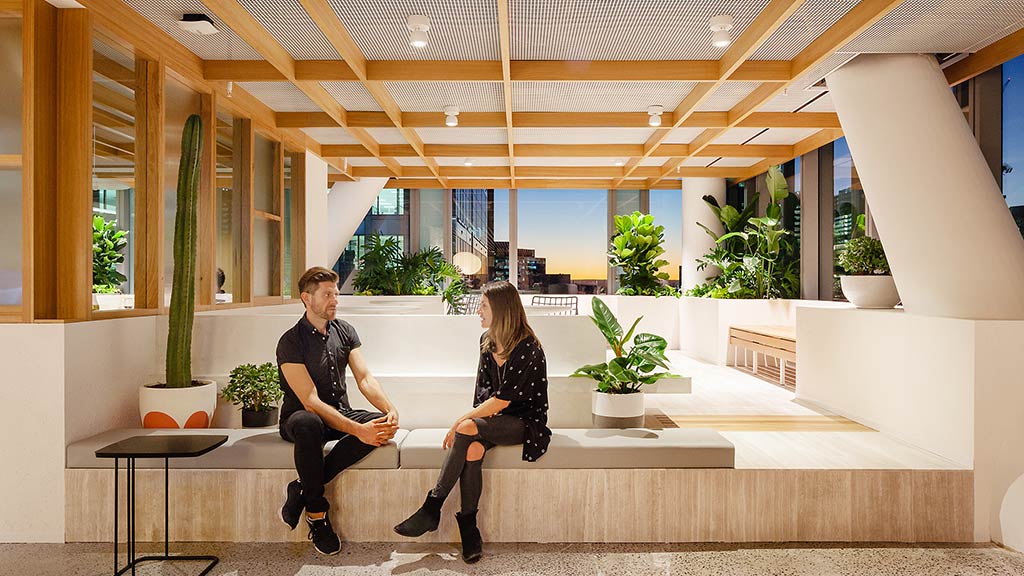 A man and woman sitting on a bench in a room with potted plants and a large window.