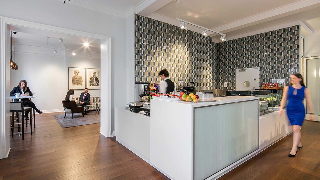 A woman in a blue dress standing in a room with a counter and people sitting at a table.