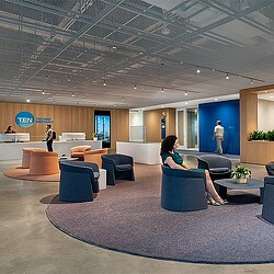A woman sitting on a chair in a large room with a man standing in the background.