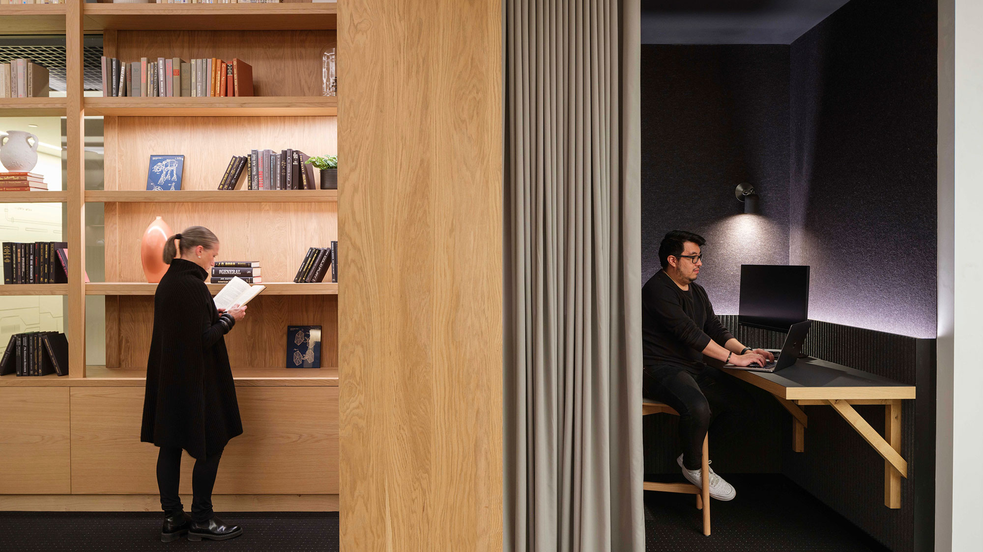 A person standing in front of a desk with a laptop and a person sitting at a desk with.
