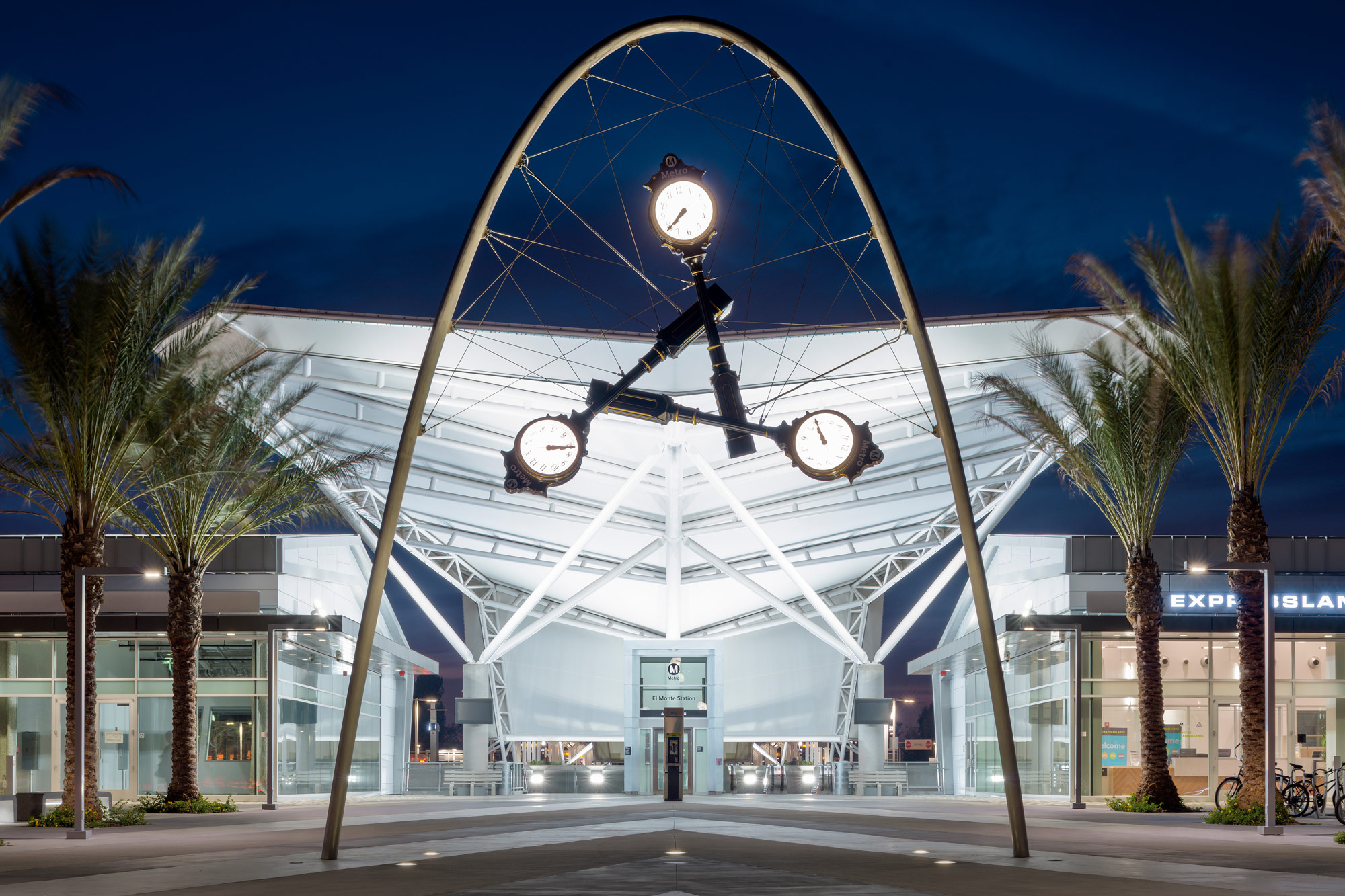 A sculpture of three suspended vintage clocks outside bus station plaza at night.