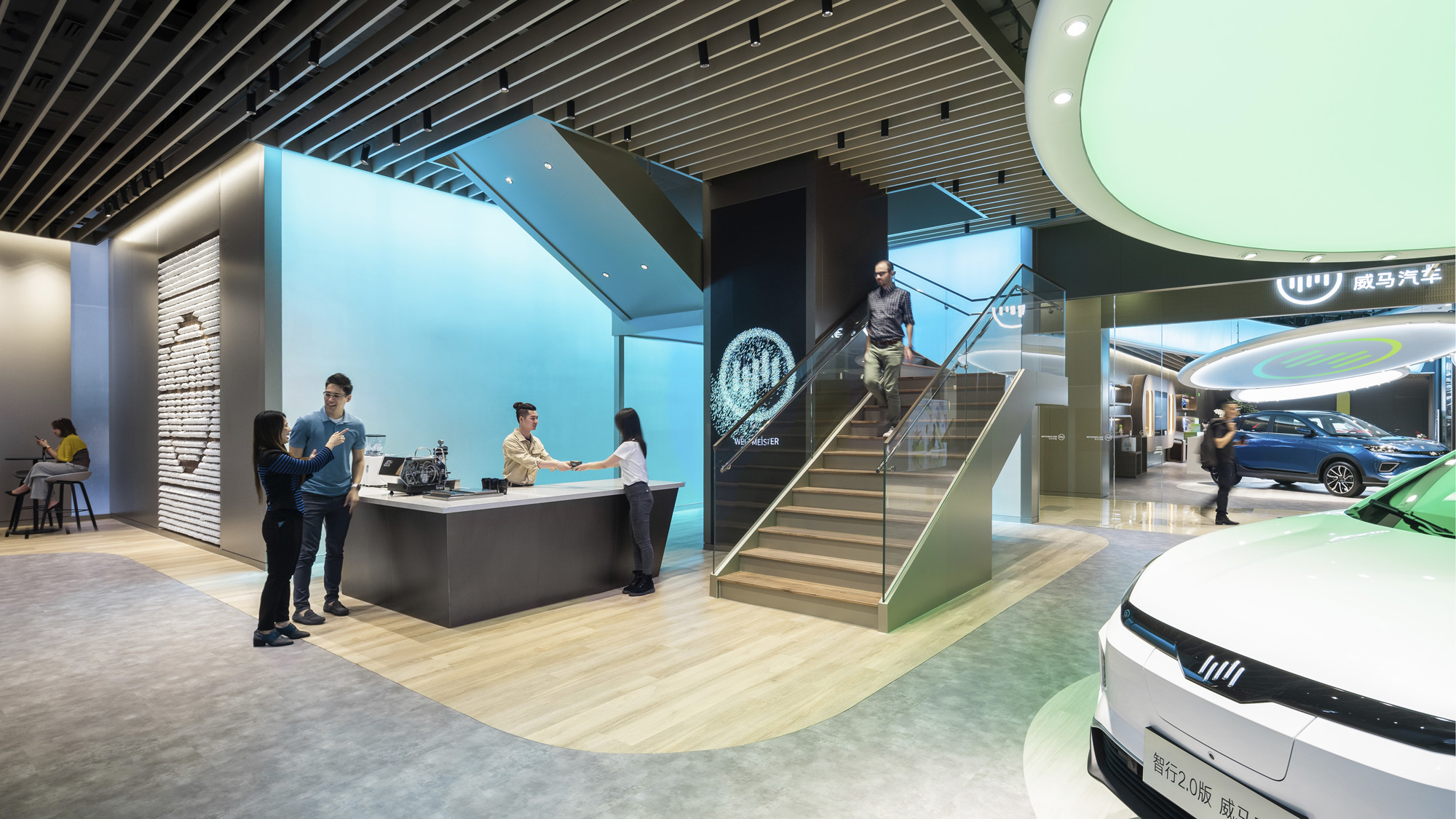 Visitors stand at a coffee bar next to a staircase and an electric vehicle showroom.