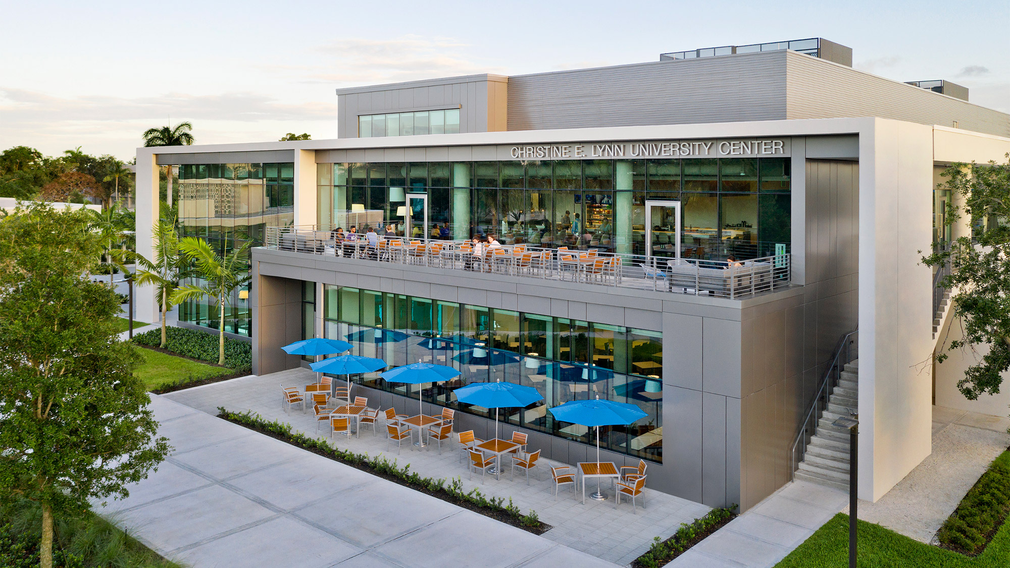 Exterior elevation photo of a two-story building with patio seating and tables with umbrellas outside and a second-story outdoor terrace with people dining.