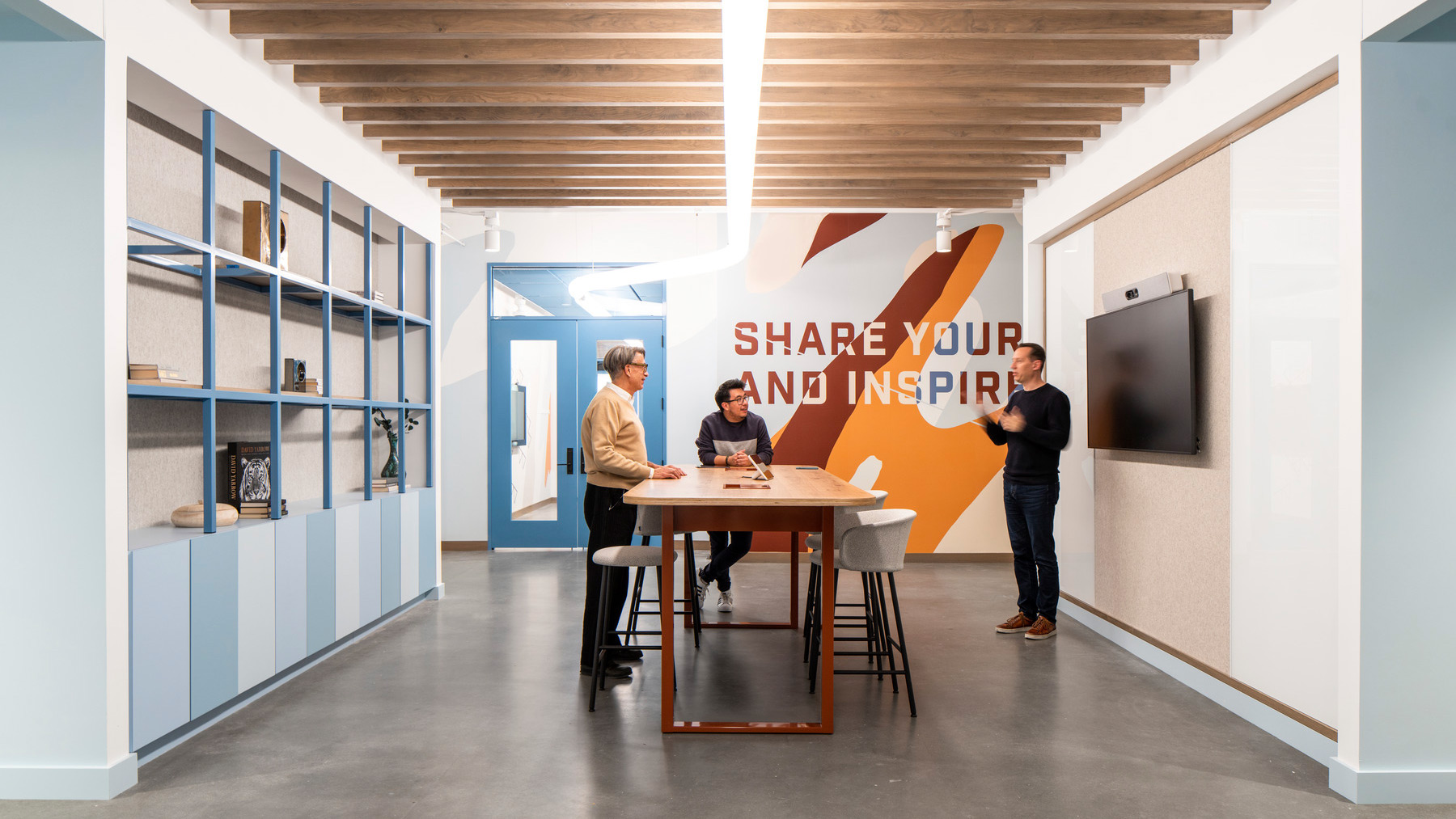 Three people are collaborating on a project while standing around a high-top conference table. An environmental graphic on the wall reads "share your inspiration".