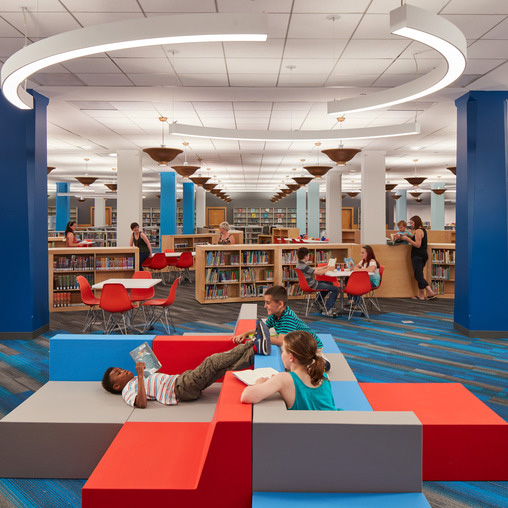 A group of people sitting on a red and blue mat in a room with tables and chairs.