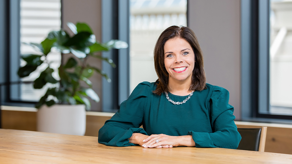 Deanne Erpelding sitting at a wooden table with a plant and windows in the background