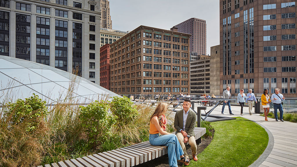 A man and woman sitting on a bench in a park with buildings.