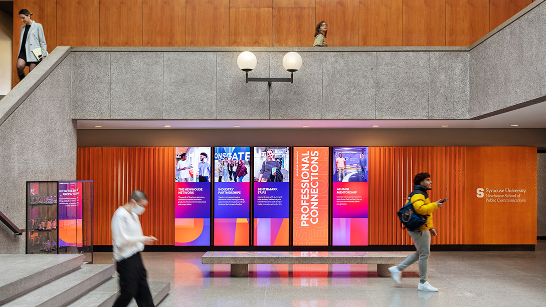 People walking in front of a wall with a large screen.