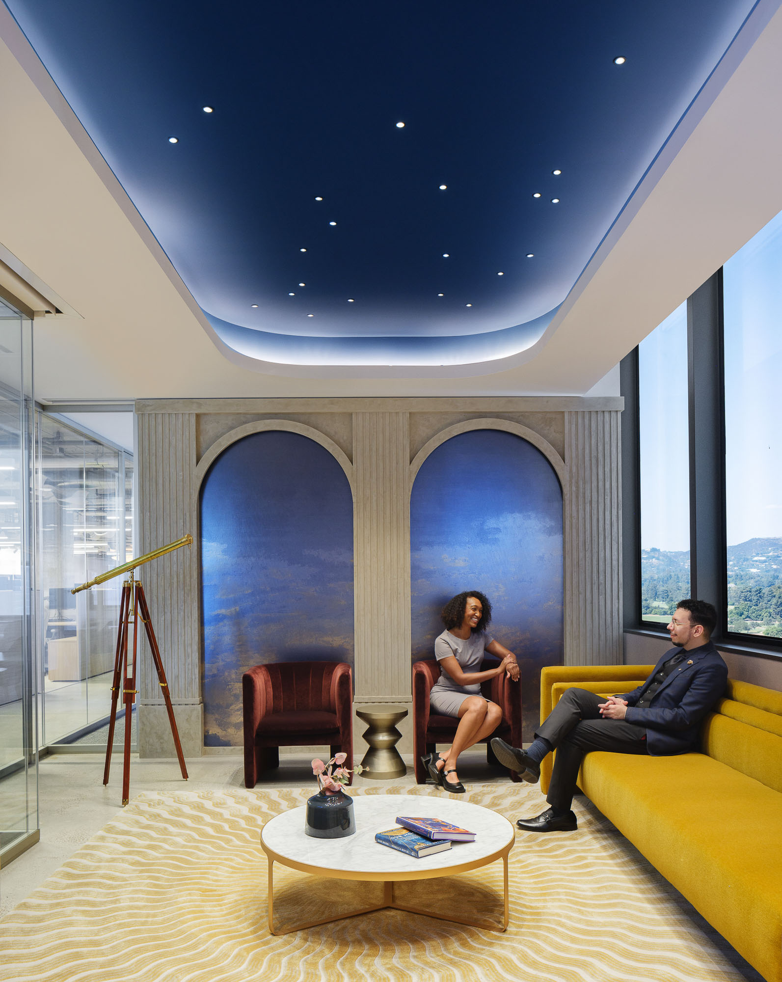 A man and woman sitting on a couch in a room with large windows.