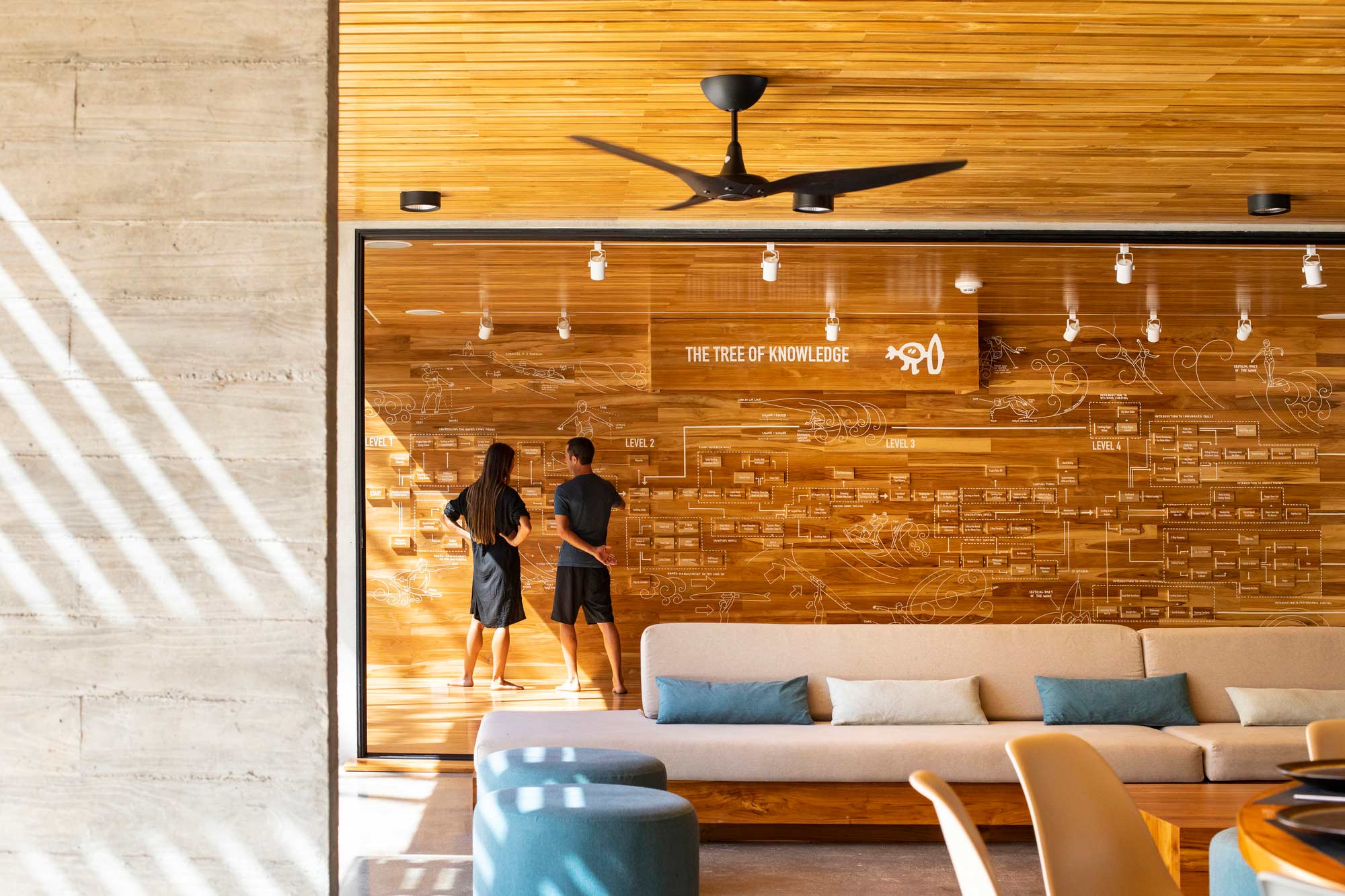 A couple of people standing in a room with a wood wall and a wood floor with a fan.
